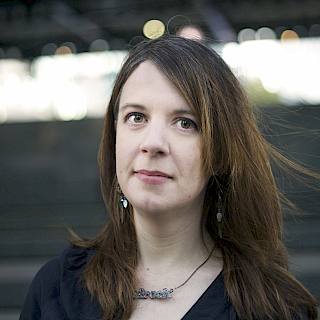 Photo of a white person with shoulder length brown hair and brown eyes. They have a necklace and a dark blue top on.