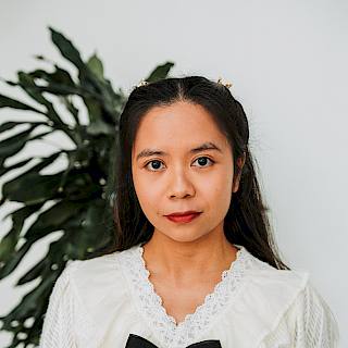 Photo of a woman with long brown hair. She wears red lipstick and a white shirt with a black bow at the collar. There is a green plant in the background.