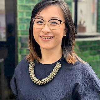 Photo of a Malaysian woman with shoulder length dark hair. She wears black framed glasses and a navy top. She also wears a large, gold necklace.