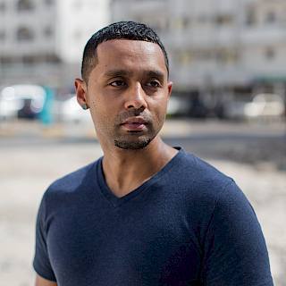 Photo shows a Brown man with short black hair. He wears a blue tshirt and there are tall buildings in the background.