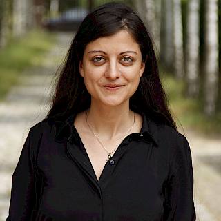Woman with long black straight hair. She wears a black shirt and a silver necklace.