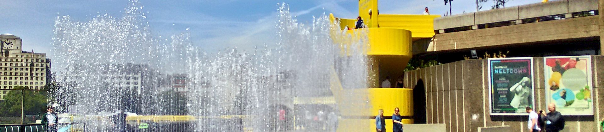 On the right is a concrete building with a bright yellow circular staircase. On the left are some waterfountains shooting into the air.