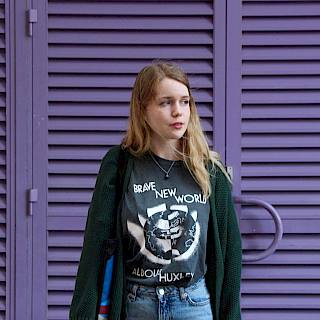 Photo of a woman with long blonde hair. She wears a tshirt with a long black cardigan and jeans. She stands in front of some purple slatted doors.