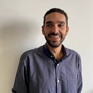 Photo of a man with short black hair. He stands against a white wall and wears a dark blue shirt.
