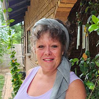 Photo of a woman with curly grey hair. It is tied up with a pale blue head scarf. She wears a lilac vest and stands in front of some vines on the side of a building.