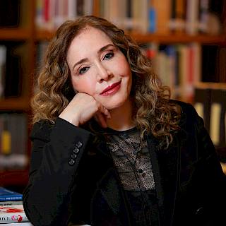 Photo of a woman in a library. She has long curly light brown hair and she is resting her head on her hand. She is wearing a black lace top and black blazer.