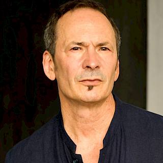 Photo of a man with short dark hair and a small goatee. He is wearing a navy blue shirt and is against a dark background.