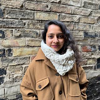 A person with long dark wavy hair stands against a brick wall. They are wearing a beige coat and white knitting scarf and are smiling into the camera.